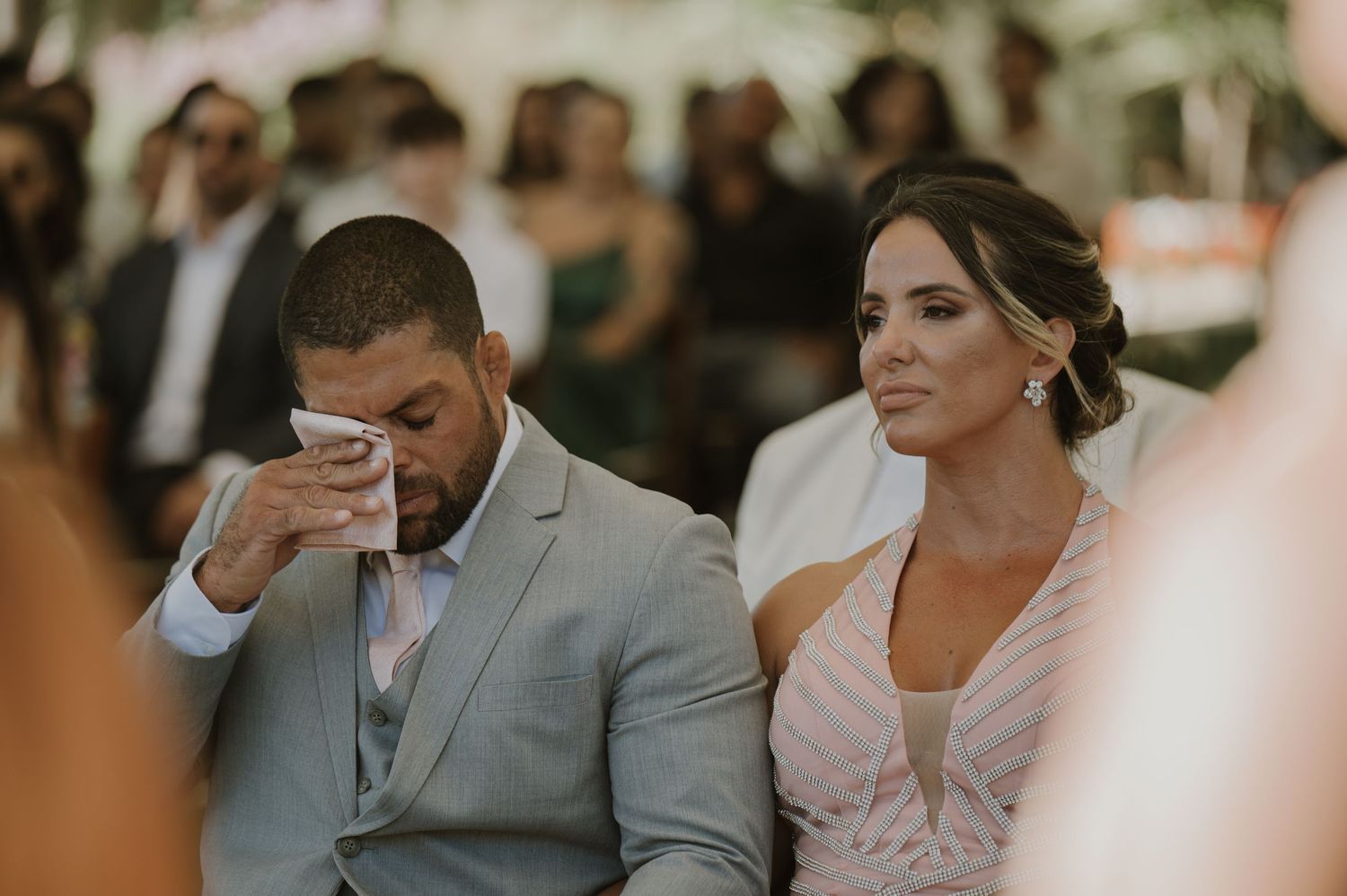 Emotional groom wipes tears while bride smiles during outdoor wedding ceremony.