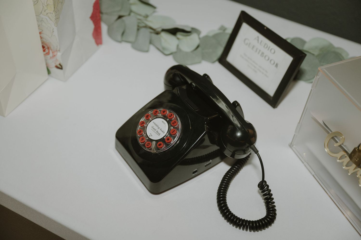 A vintage black rotary dial telephone sits on a white table next to a framed sign.