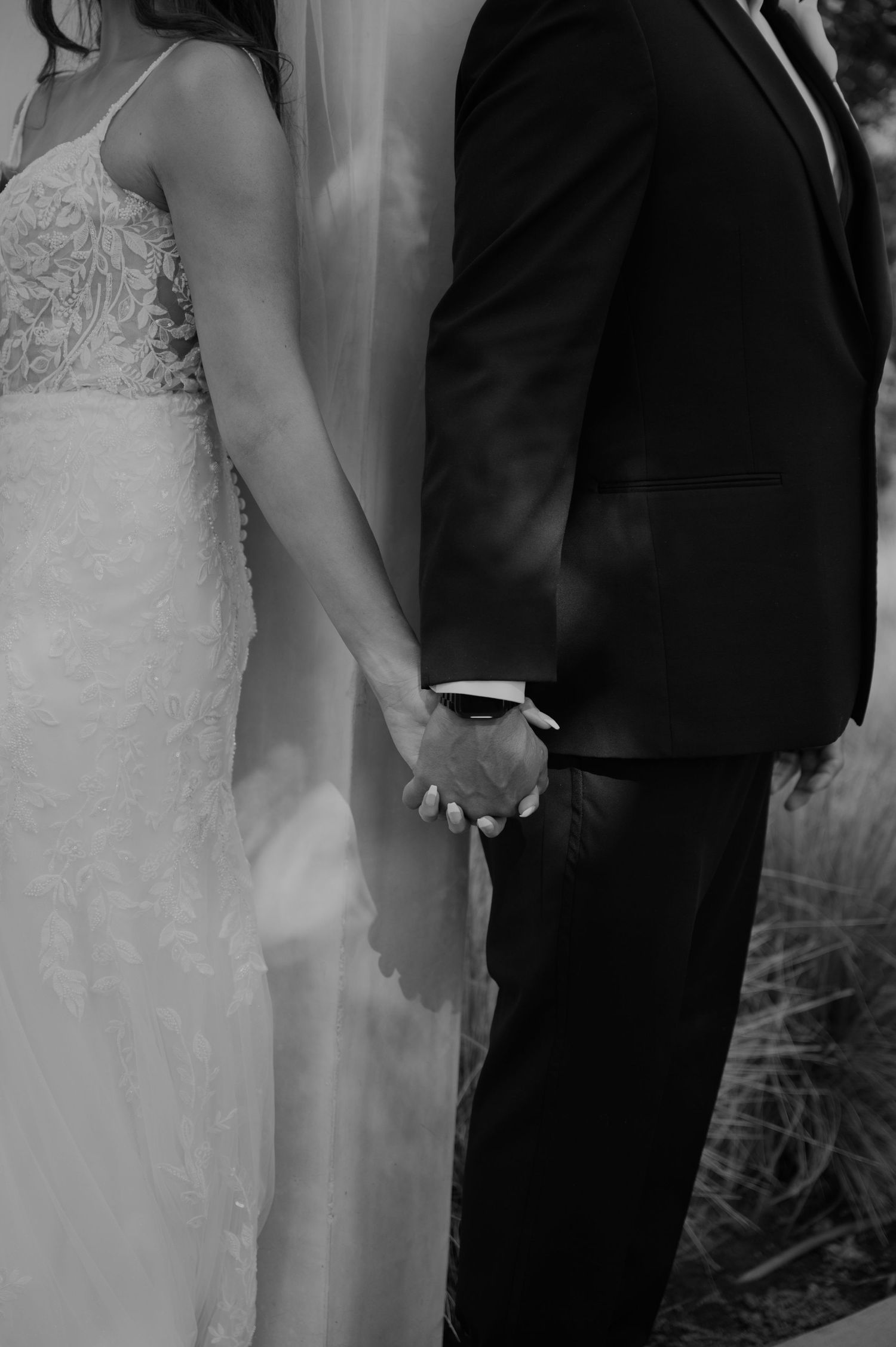 Black and white photo of bride and groom holding hands, bride in lace gown and groom in dark suit.