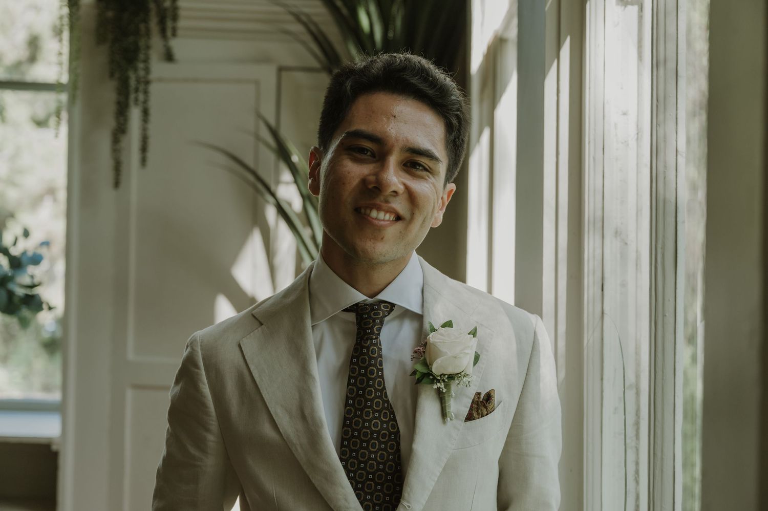 Smiling groom in white suit with floral boutonniere and polka dot tie standing by a bright window.