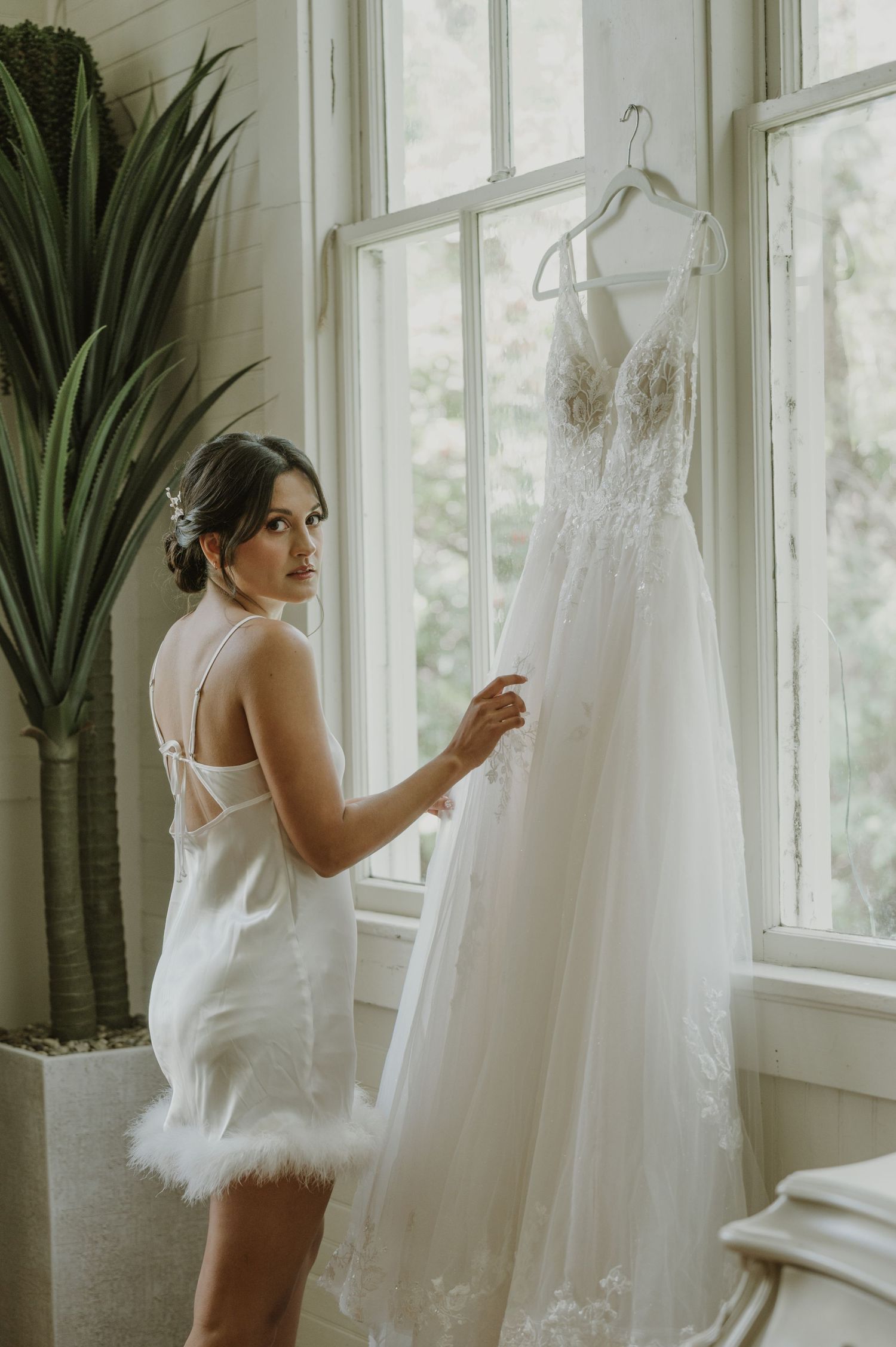 Bride in white mini dress with feather hem admires her wedding gown hanging by a sunlit window.