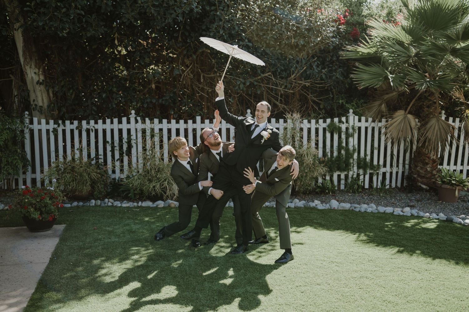 Groomsmen in black suits playfully pose with a white parasol on a green lawn at an outdoor wedding.