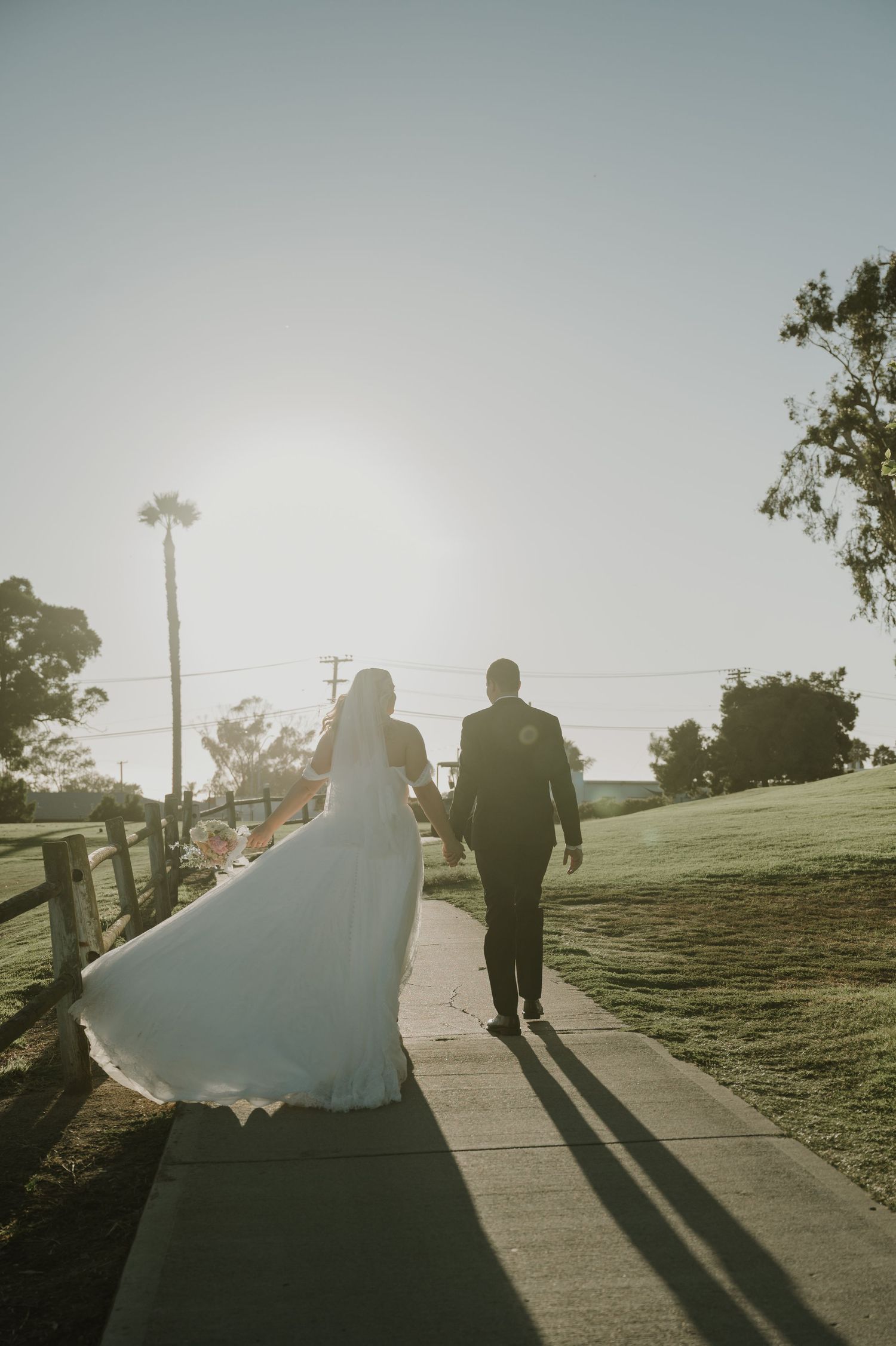 Bride and groom walking hand in hand down a sunlit path on their wedding day.