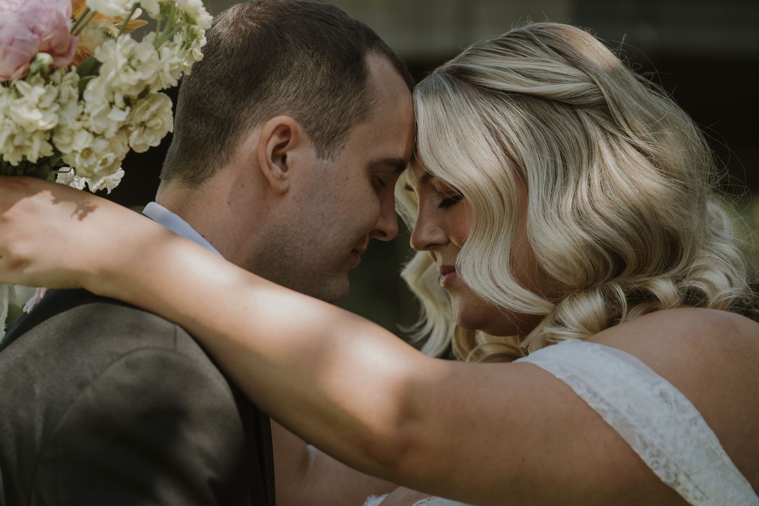 A bride and groom share an intimate moment, touching foreheads, with the bride wearing a floral hair piece on their wedding day.