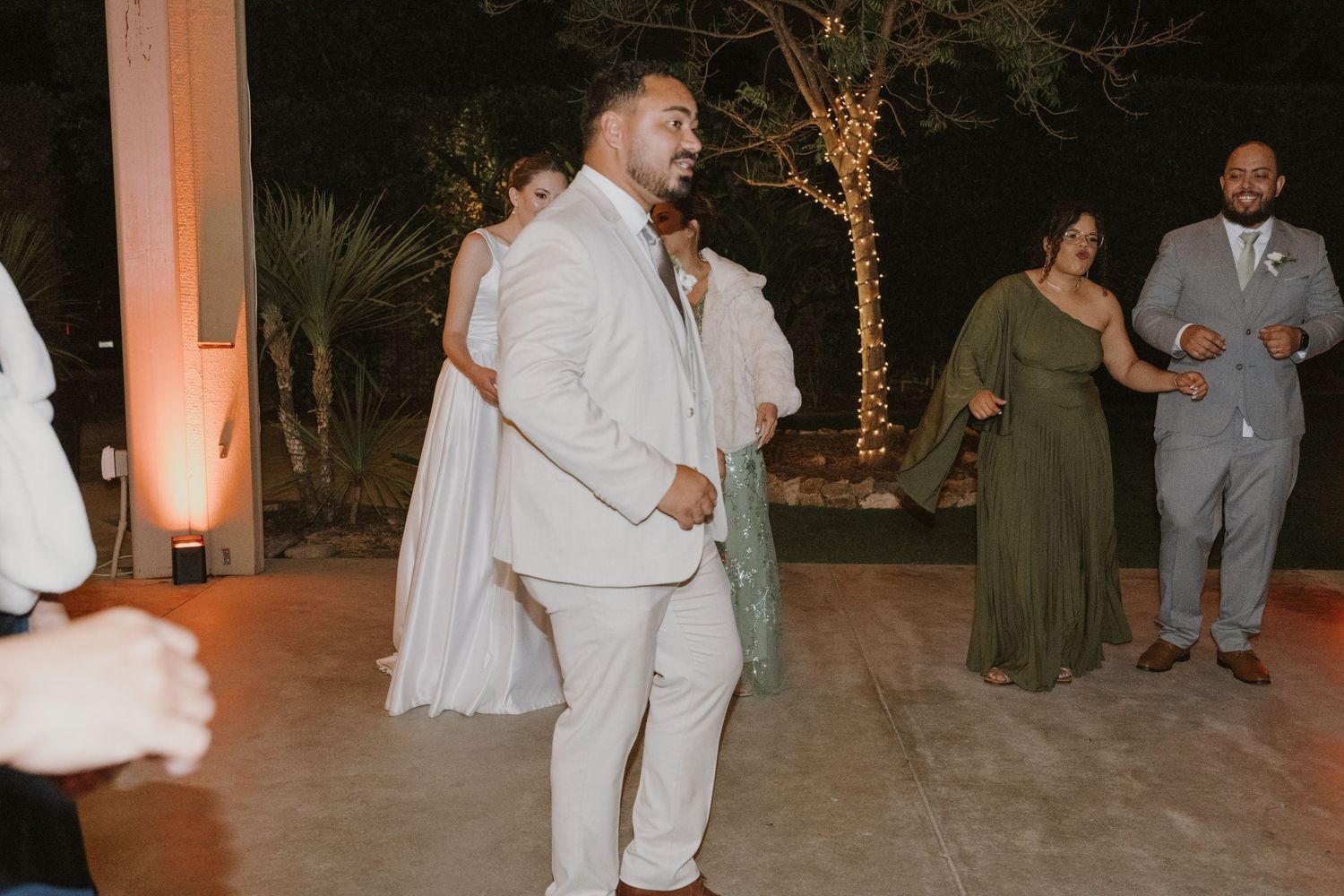 Groom in white suit dancing at outdoor wedding reception with guests in formal attire at night.