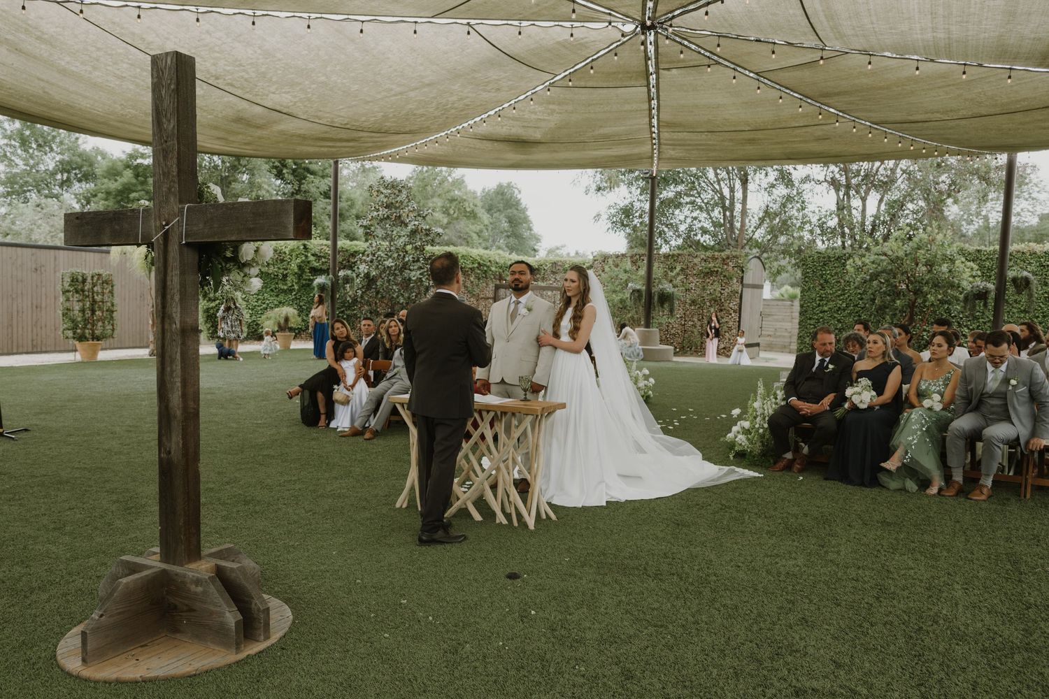 Bride and groom exchange vows at outdoor wedding ceremony beneath a canopy with cross decoration.
