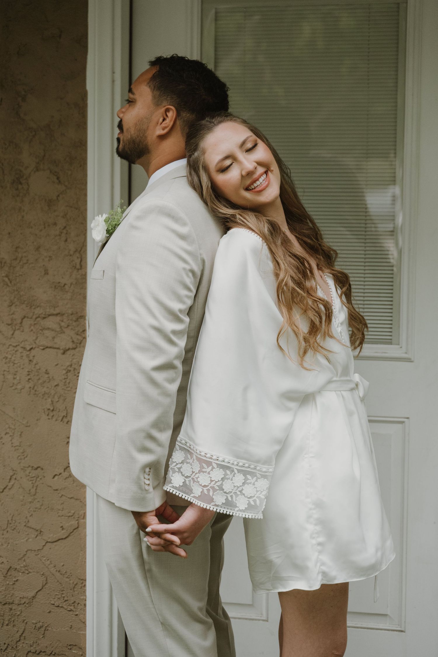 Bride in white satin robe holds hands with groom in beige suit, smiling back-to-back in a first look moment.