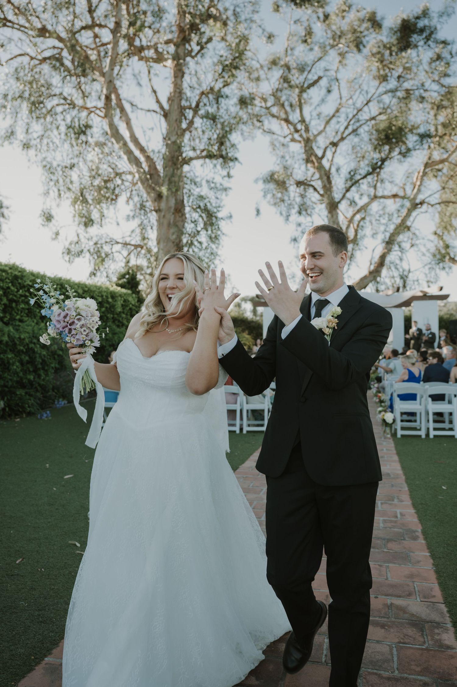 Beaming newlyweds show off wedding rings while walking down aisle after outdoor garden wedding ceremony.