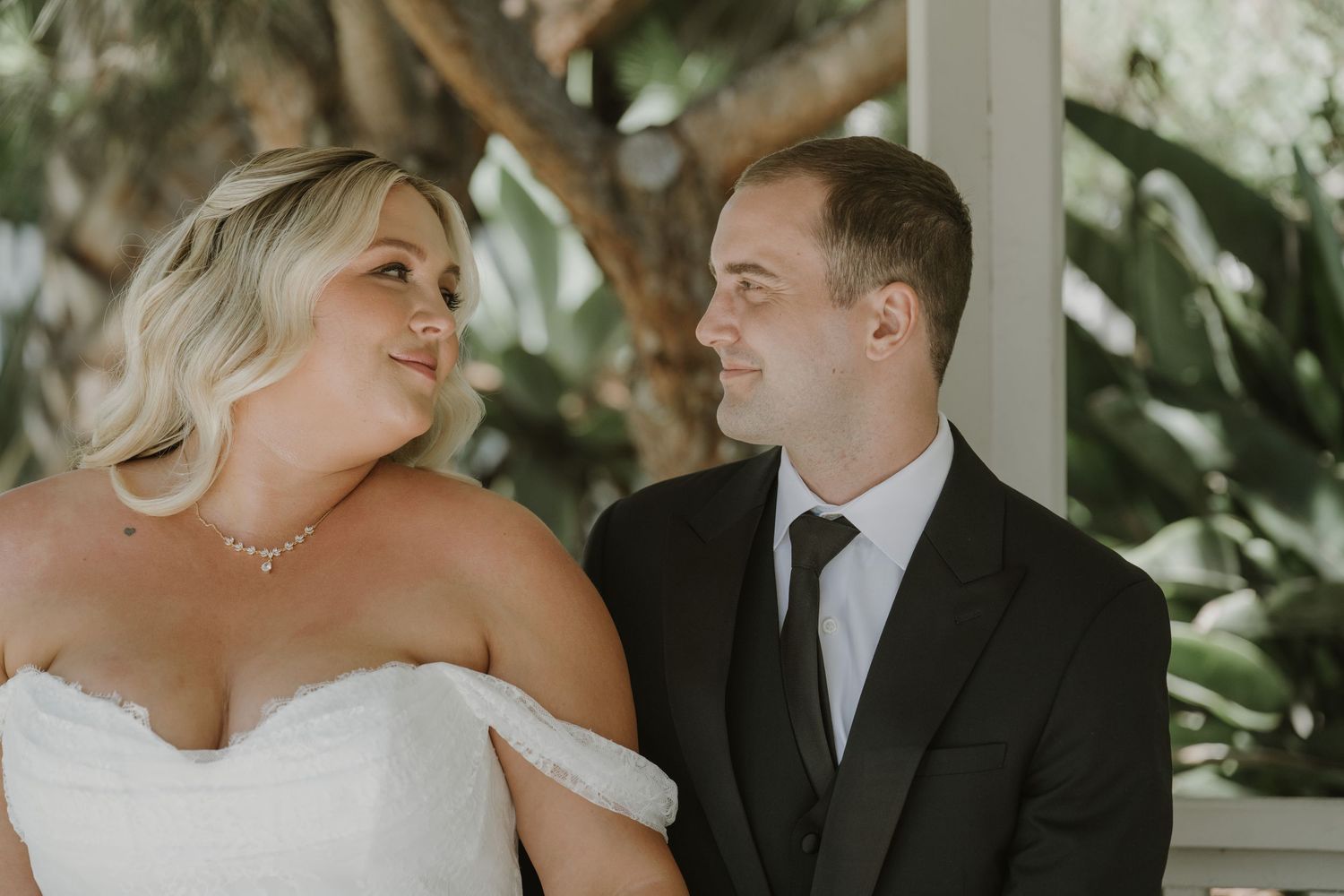 Bride and groom share a loving glance, dressed in wedding attire with lush greenery in background.