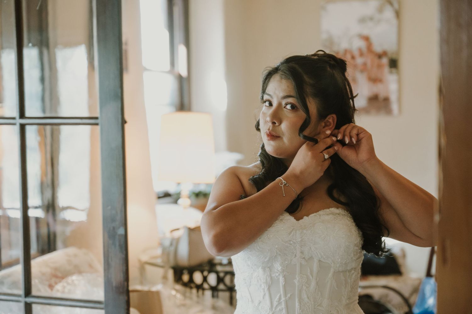 Bride in white lace gown putting on earrings while getting ready in a softly lit room.