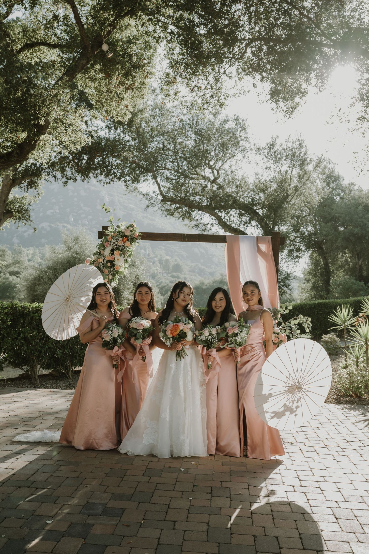 Bride and four bridesmaids in blush pink gowns holding white parasols and bouquets at outdoor wedding.