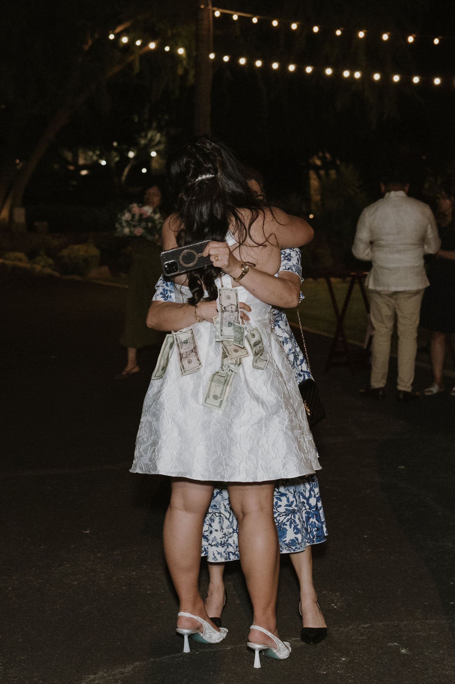 Two women in floral dresses embracing on outdoor wedding dance floor under string lights at night.