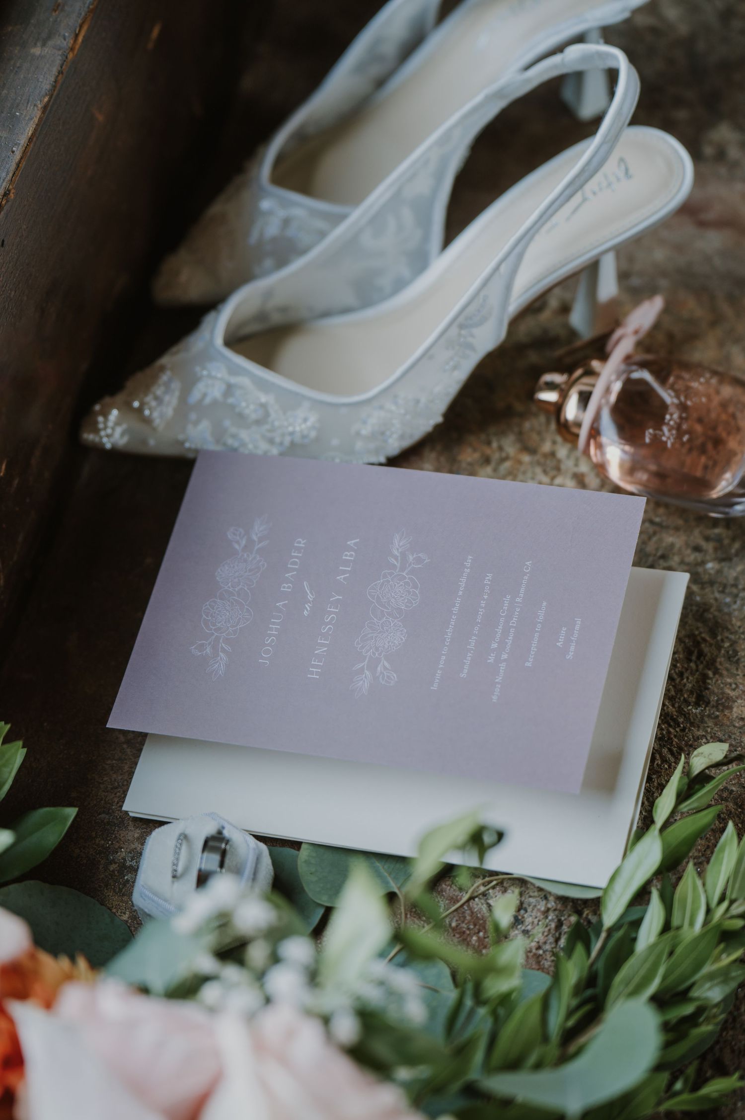 Elegant wedding flat lay featuring lace heels, lavender invitation, floral bouquet, and perfume bottle on rustic wood surface.