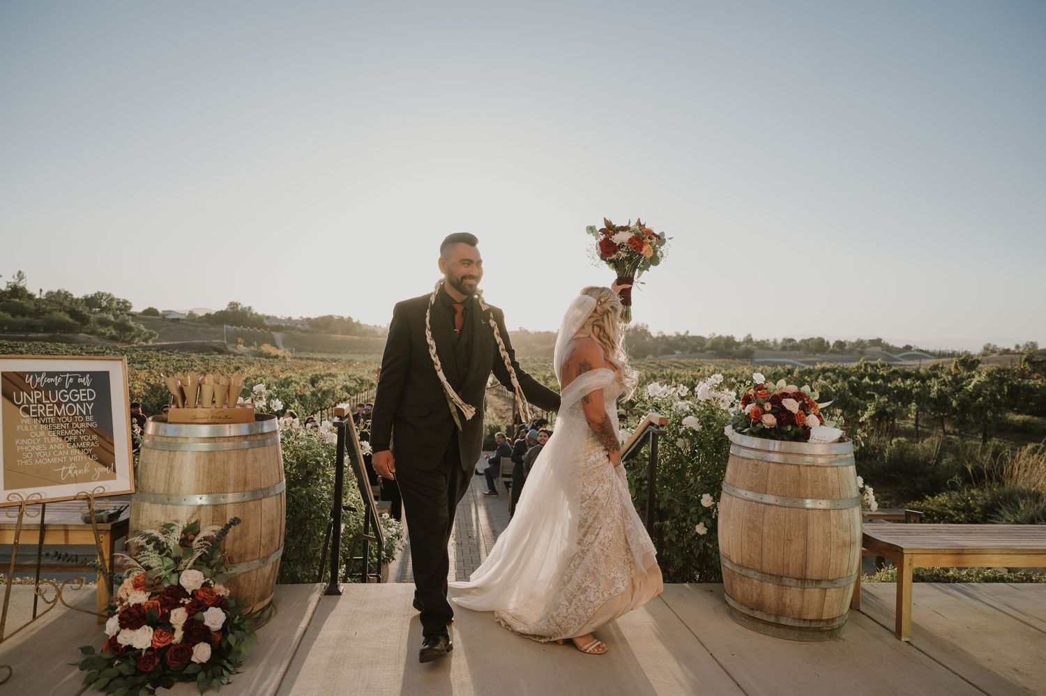 Romantic wedding ceremony setup at a scenic vineyard with wine barrels and floral arrangements at sunset.
