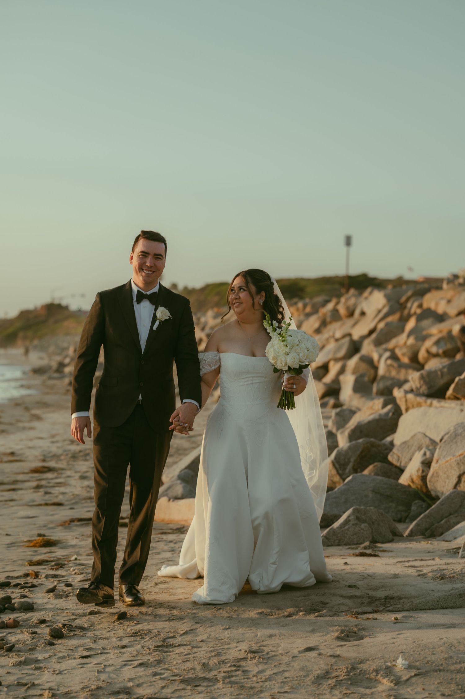 Bride and groom walking hand-in-hand on a sandy beach at sunset, bride holding white bouquet in elegant wedding attire.