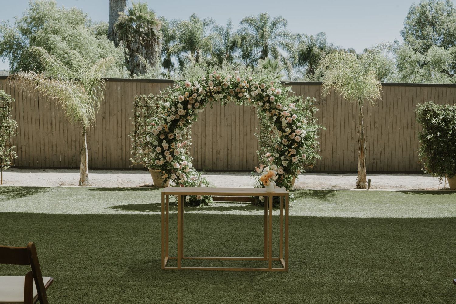 Circular floral wedding arch with white and pink blooms on green lawn, framed by palm trees and bamboo fence.