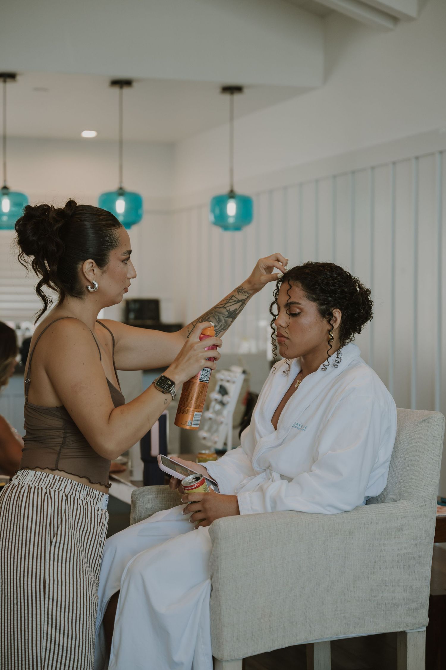 Hair stylist applying product to bride's curly updo while she sits in white robe before wedding ceremony.