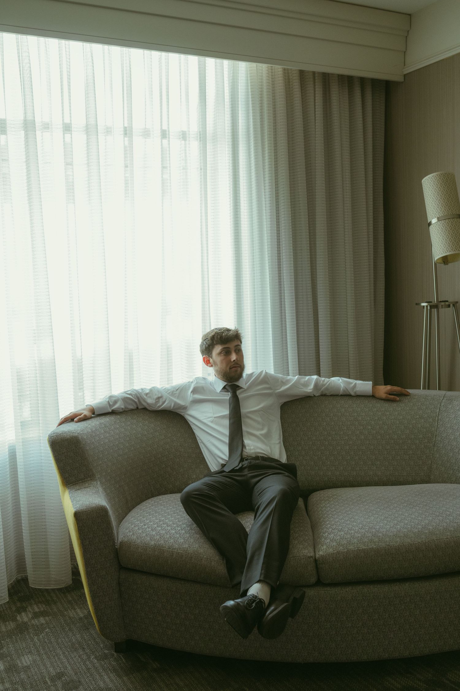 A businessman in a white shirt and tie relaxes on a sofa with arms spread wide in a modern hotel room.