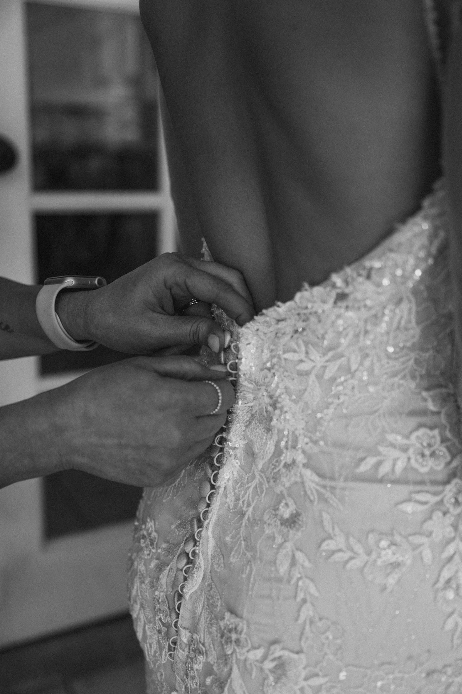 Black and white photo of hands fastening buttons on a lace wedding dress with an open back.
