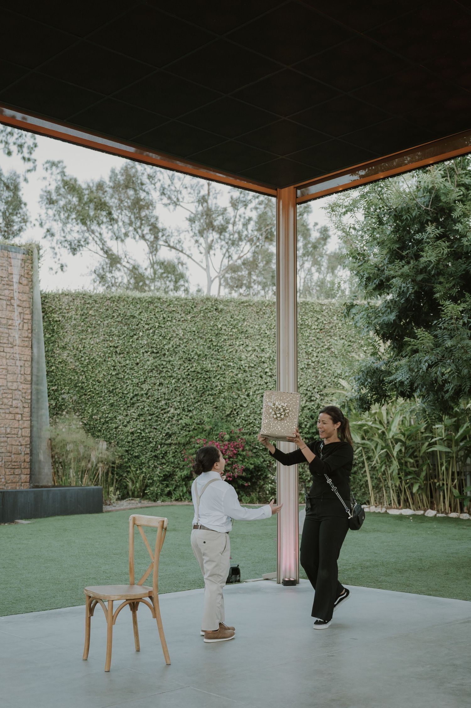 Woman in black outfit holds a decorative item while a child in white kneels on one knee under an outdoor pavilion.