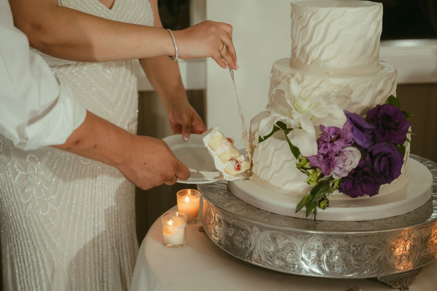 Bride and groom cut their elegant white three-tier wedding cake adorned with purple and white flowers.