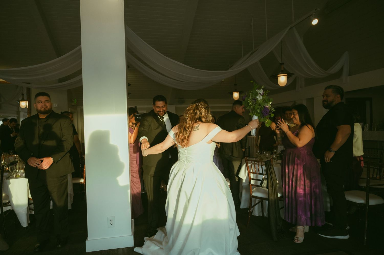 Bride in white gown dances joyfully with guests during lively indoor wedding reception celebration.