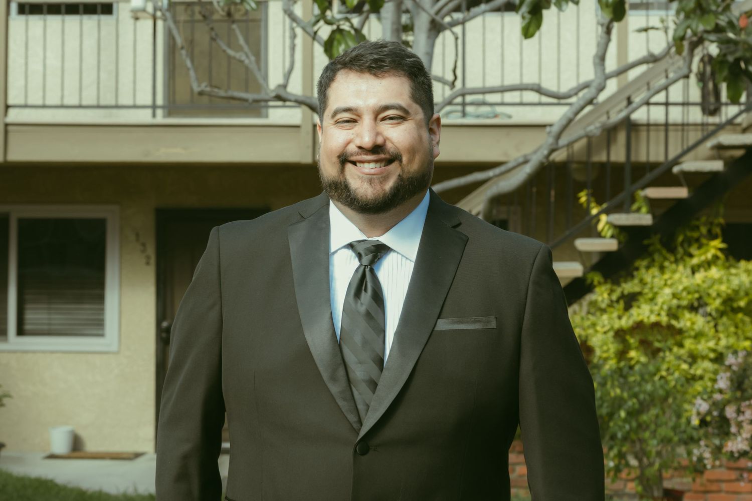 Smiling bearded man in a brown tuxedo with tie posing outdoors near a building.