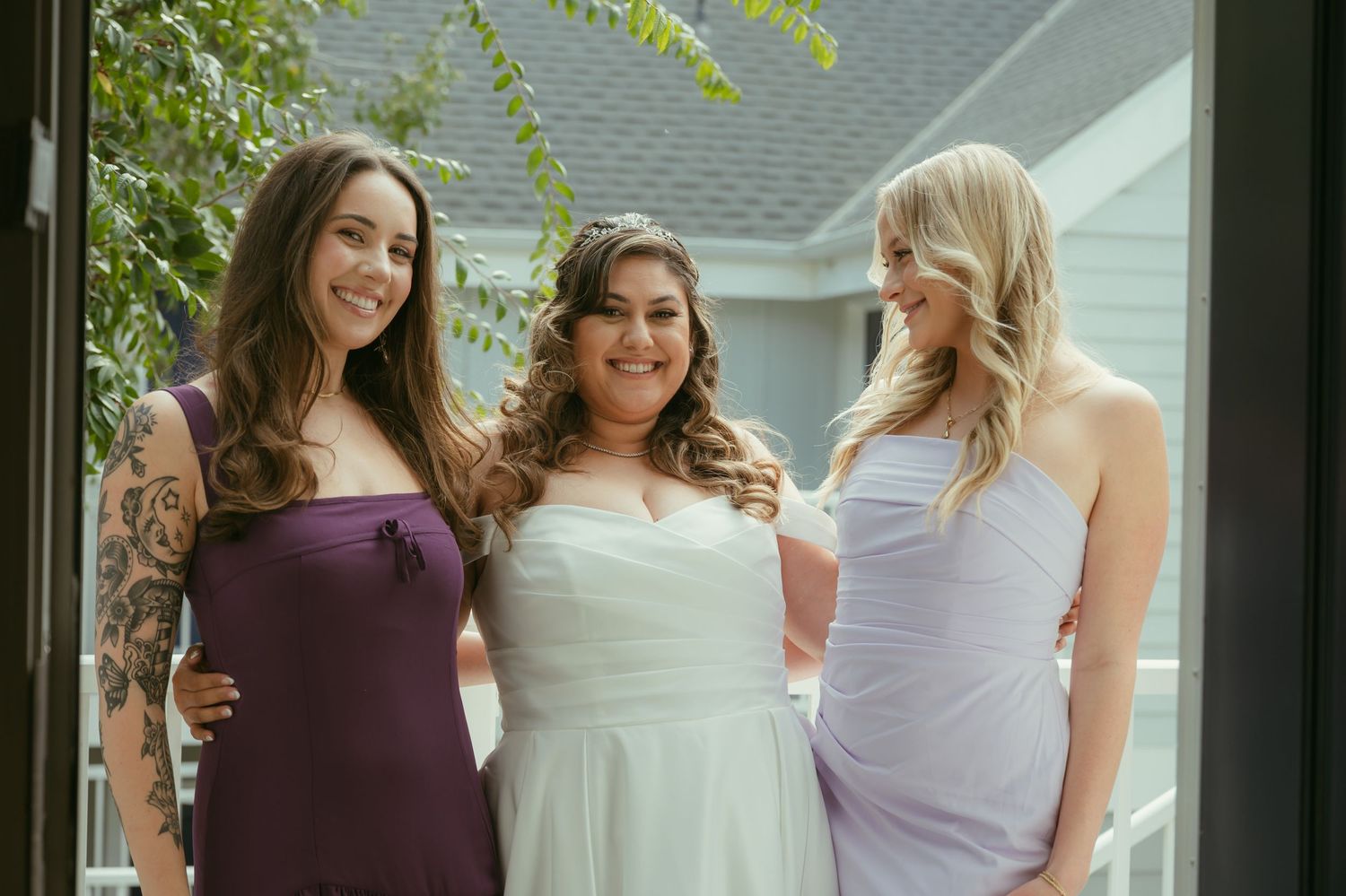 Bride in white gown poses with two bridesmaids in purple dresses, smiling outdoors near a doorway.