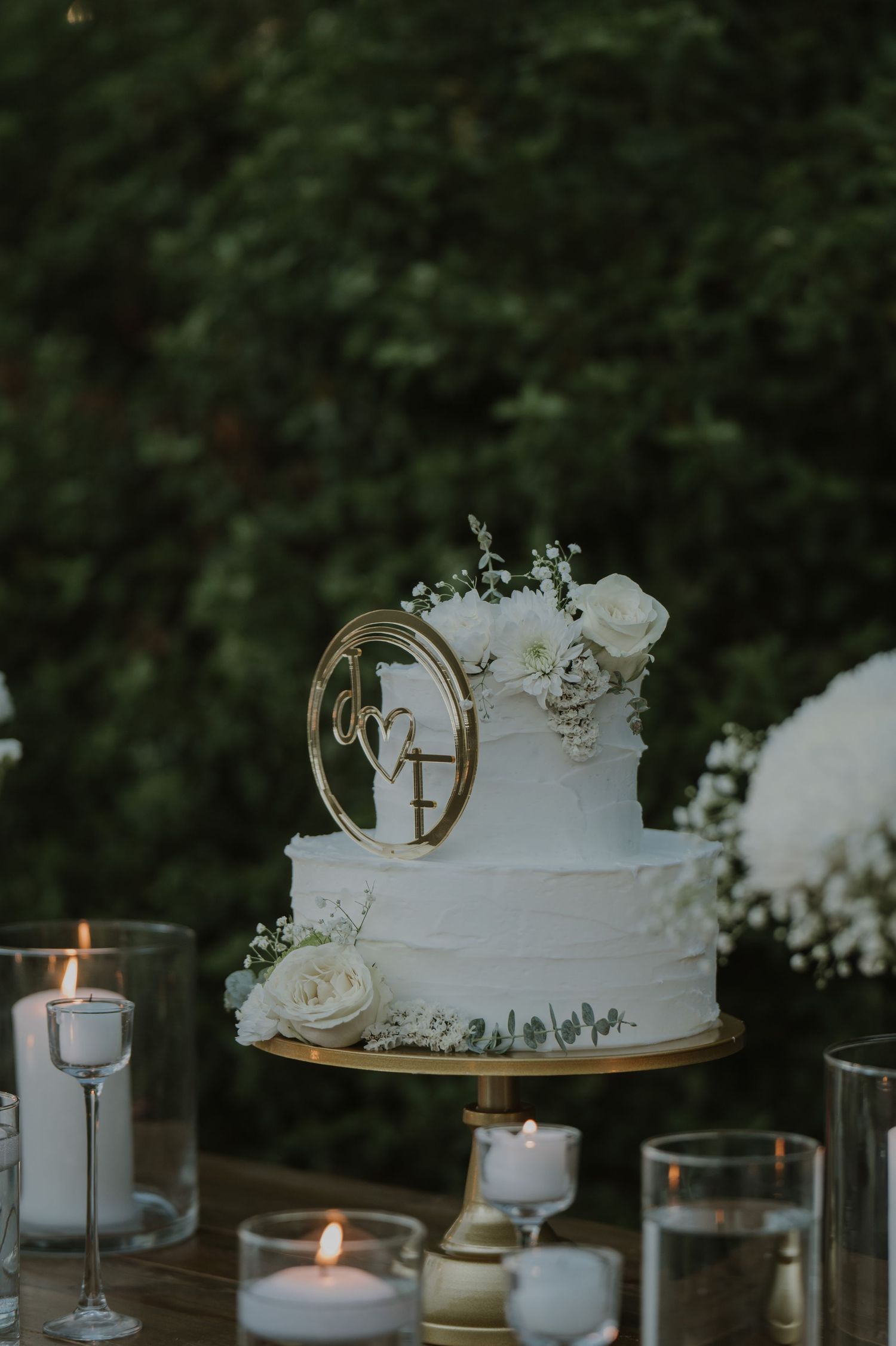 Elegant two-tier white wedding cake with gold monogram topper and white roses on a gold stand surrounded by candles.