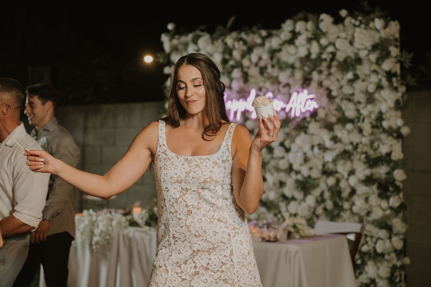 Bride in lace wedding dress dancing elegantly in front of floral backdrop with neon sign at night.