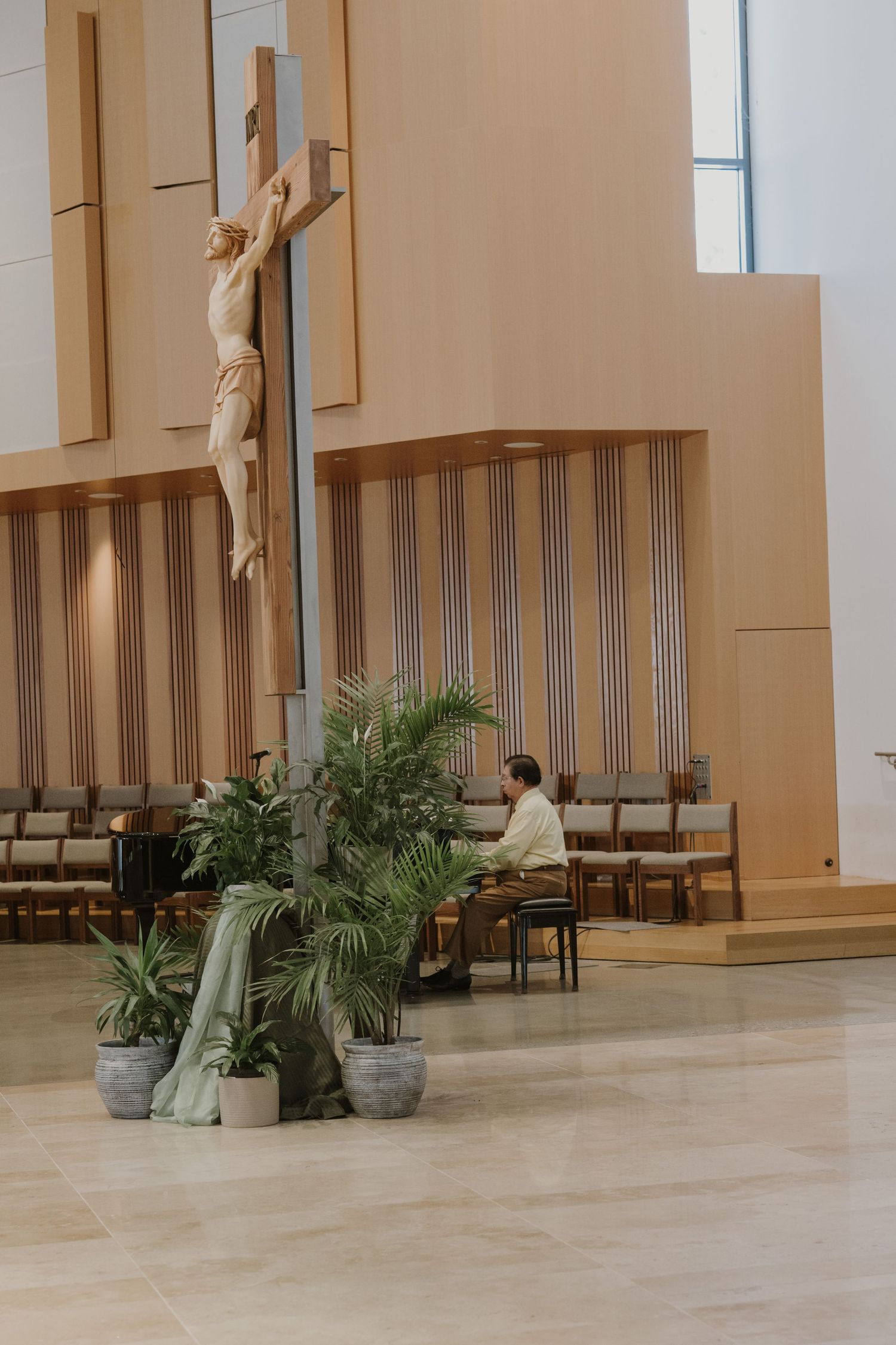 Person praying alone in a modern Catholic church with a large crucifix and tropical plants in the foreground.