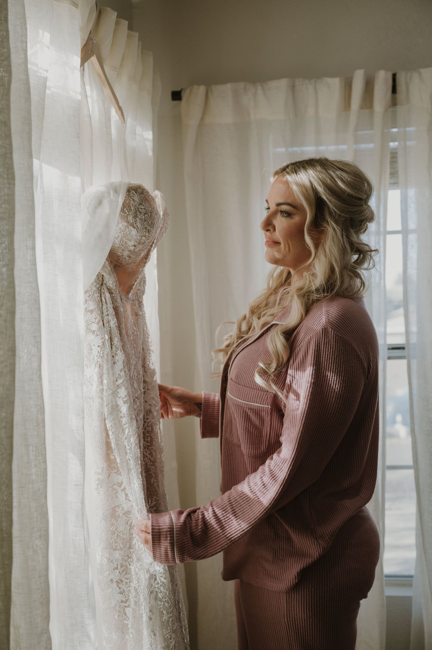 Elegant wedding dress hanging by sheer curtains in natural window light.