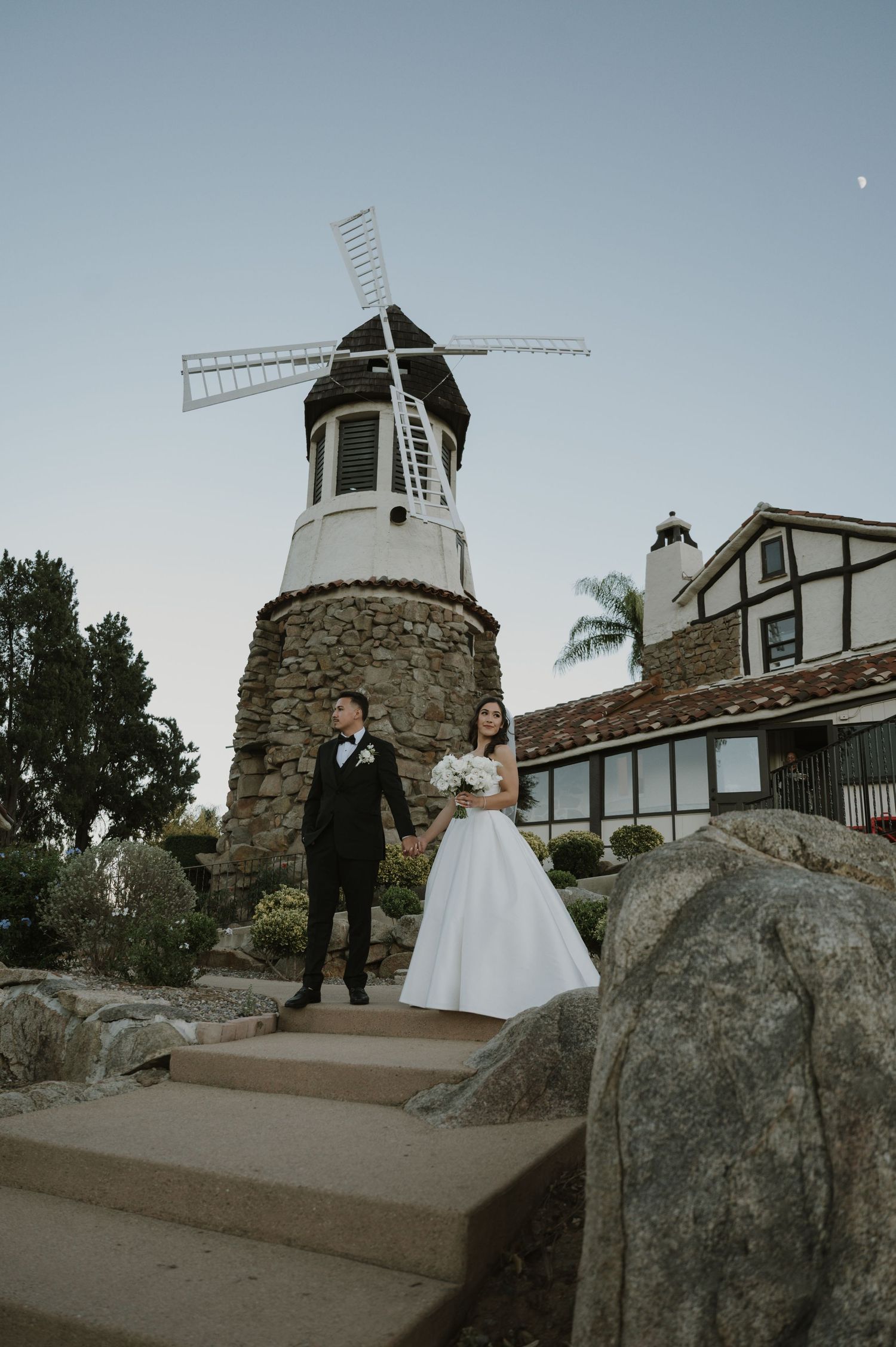 Wedding couple poses in front of white windmill and stone building at sunset.