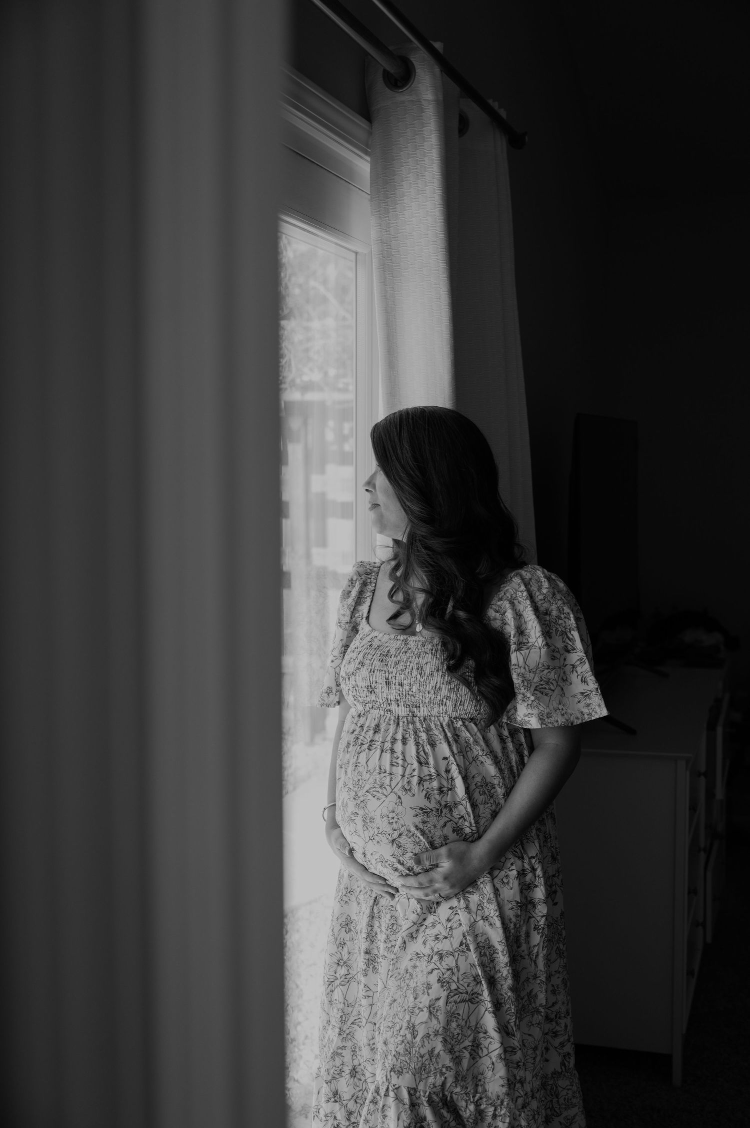 A silhouetted figure in a floral dress stands by a window during a maternity photoshoot.
