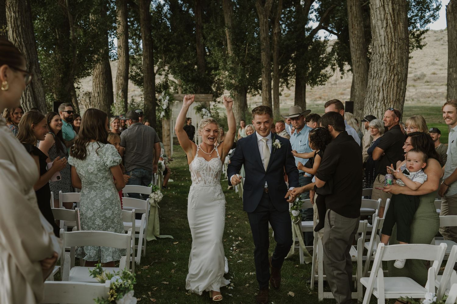 Guests mingle during a wedding reception held outdoors among tall trees and white chairs.