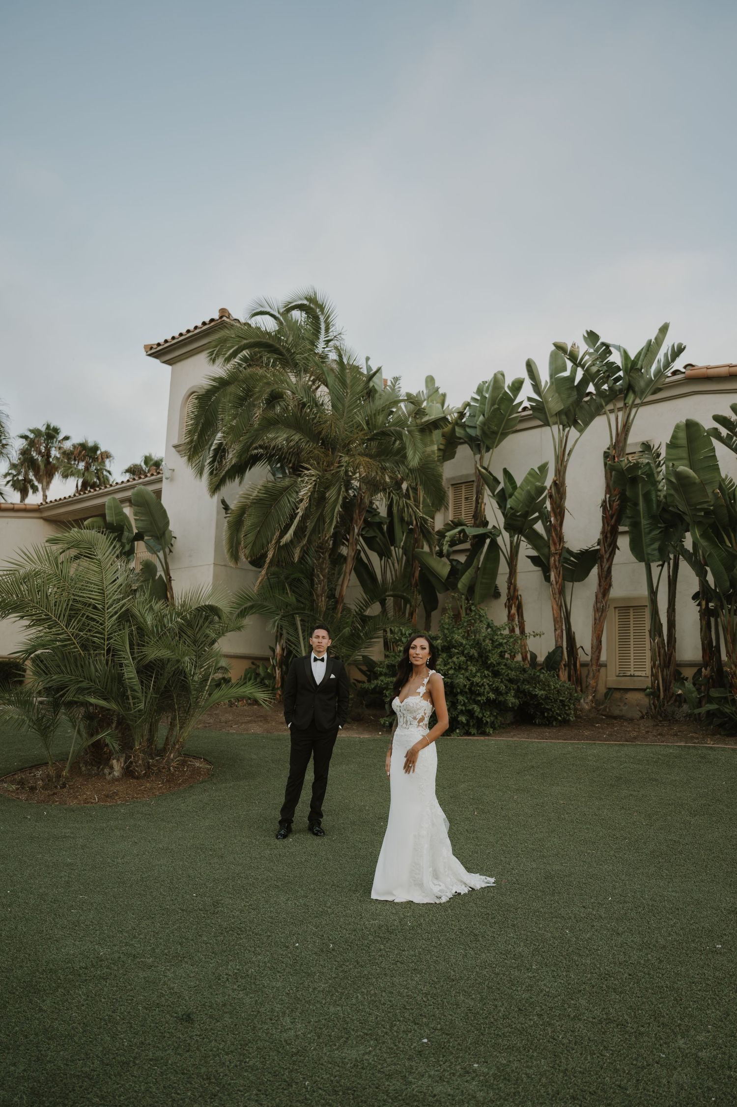 A wide-angle wedding photo showing a couple on a manicured lawn with Spanish-style architecture.
