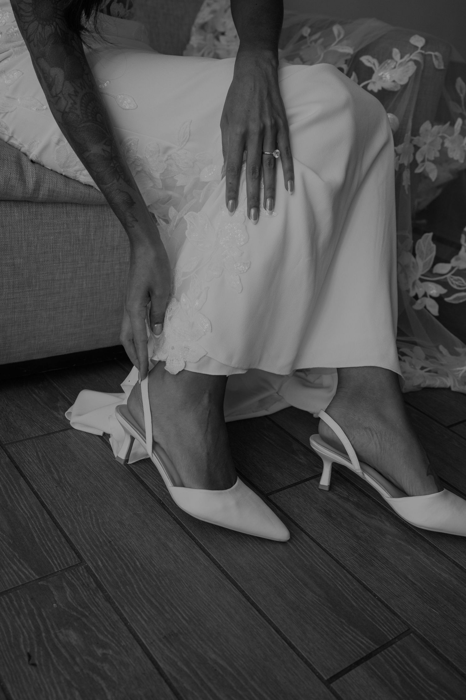 Black and white photo of wedding shoes and dress hem on hardwood floor.