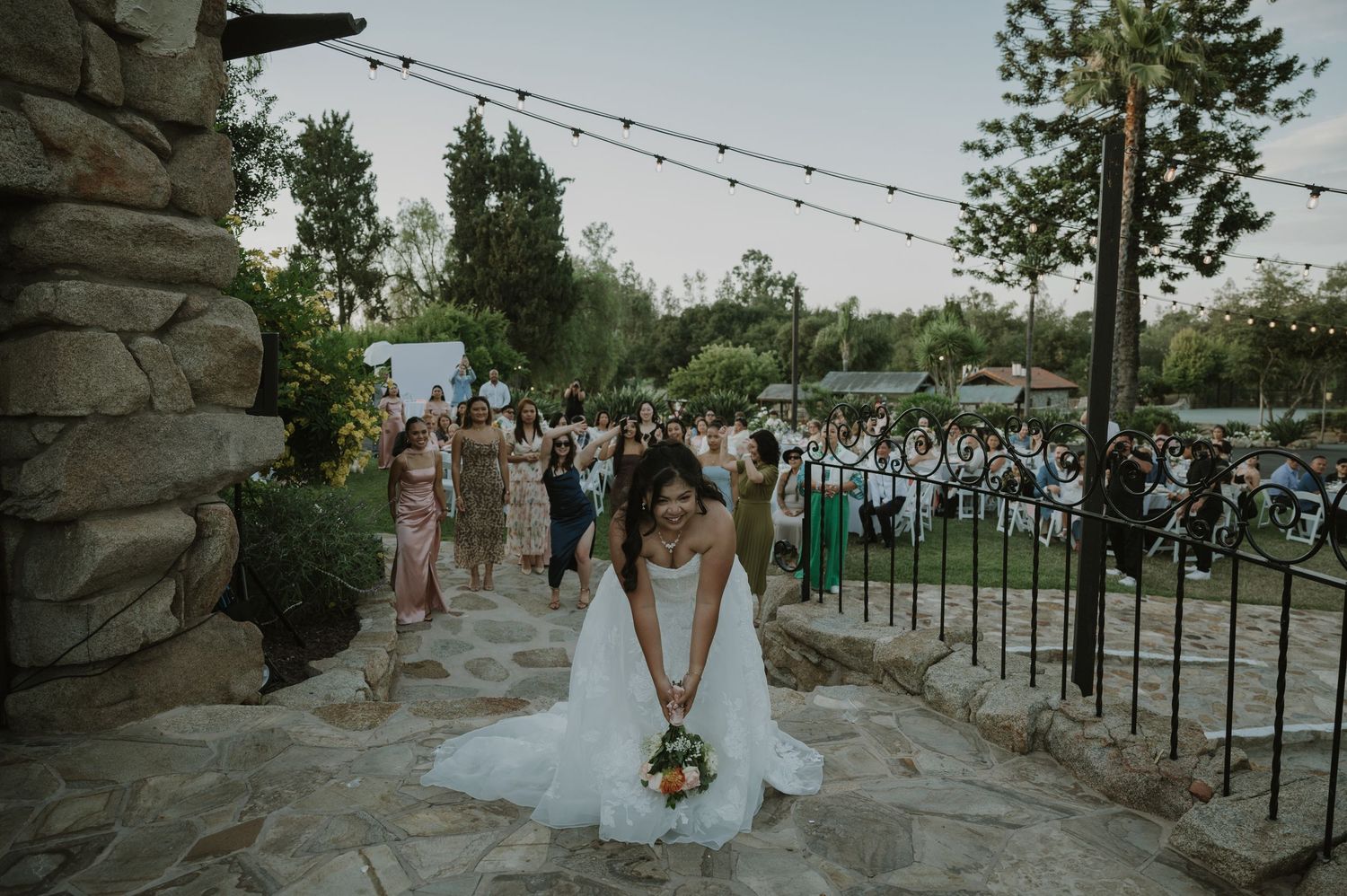 Bride in white gown walks down stone path at outdoor wedding ceremony with guests seated in background.