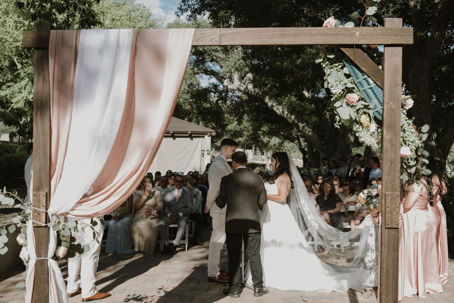 Bride and groom exchange vows under a rustic wooden arch with blush pink draping at an outdoor wedding ceremony.