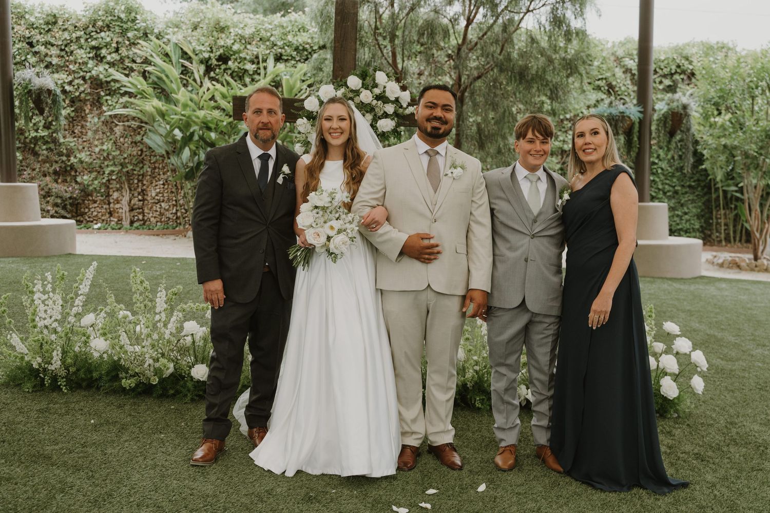 Wedding party poses outdoors with bride and groom in center, flanked by family members in formal attire.
