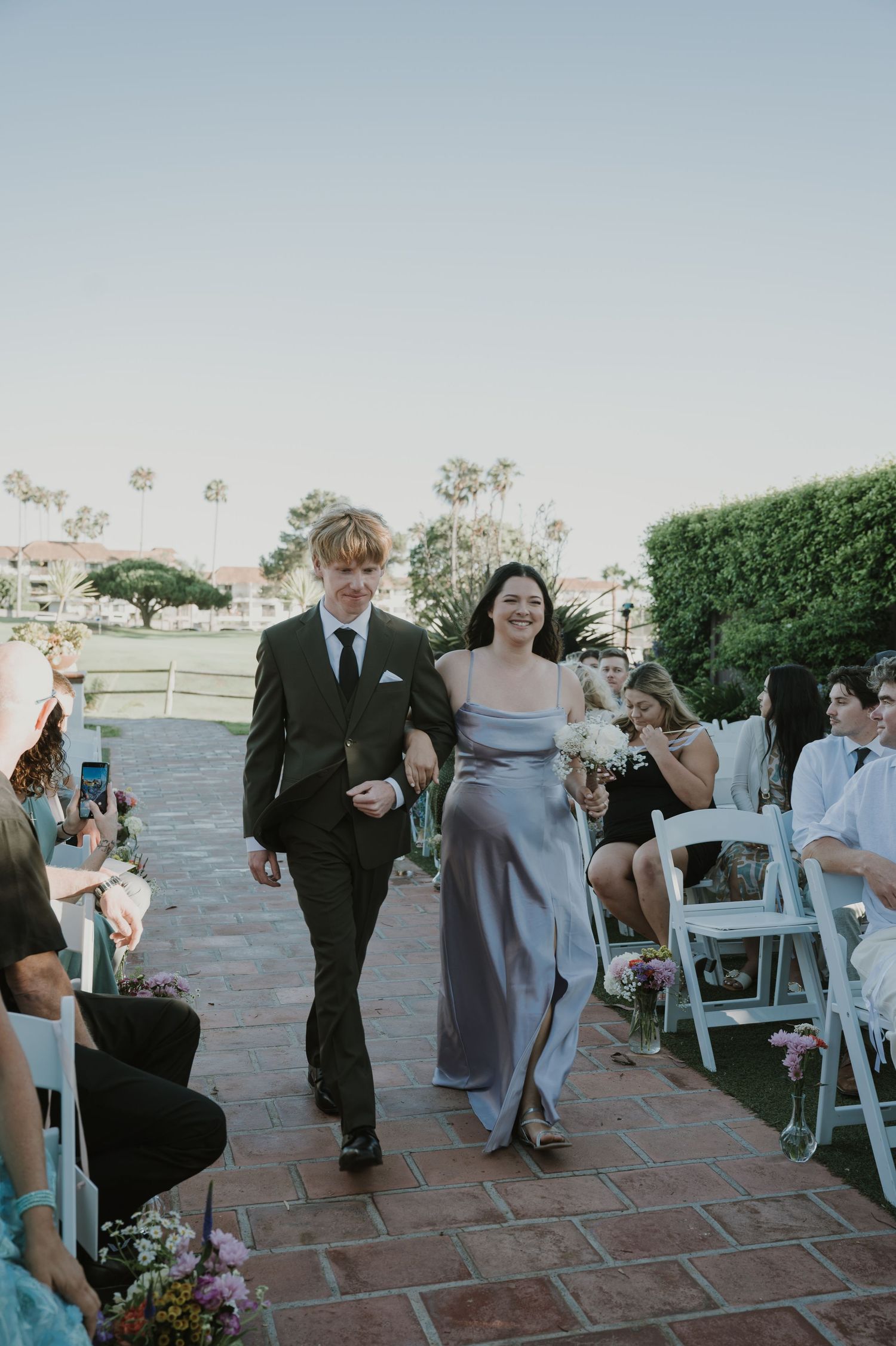 Groomsman in dark suit escorts smiling bridesmaid in silver gown down brick wedding ceremony aisle.