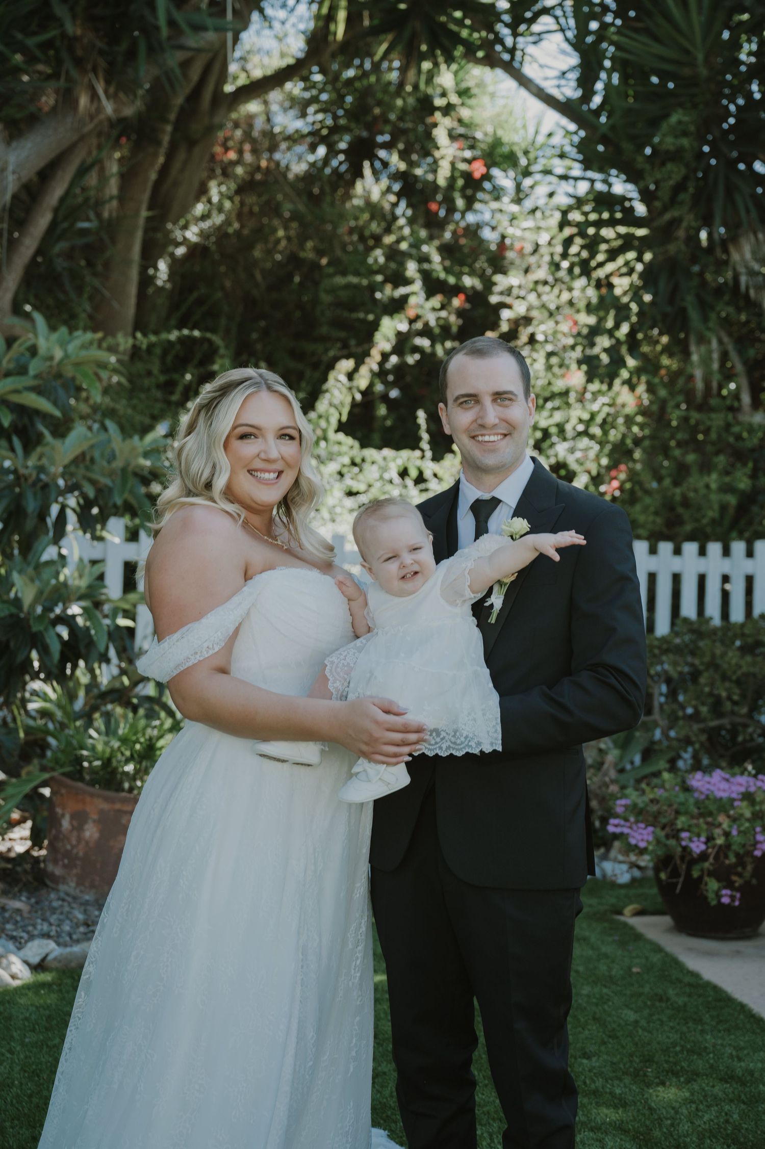 Bride and groom smiling in a garden, holding a baby in a white dress on their wedding day.