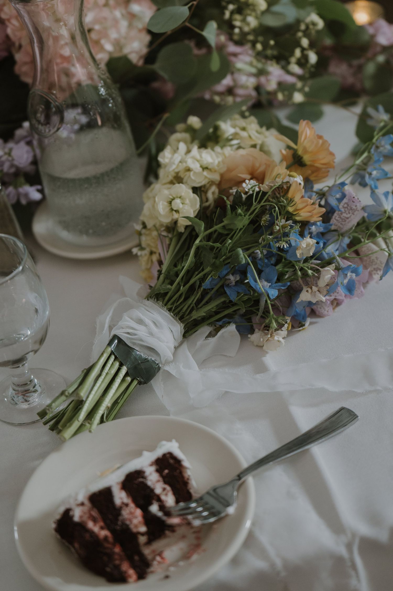 Wedding table with chocolate layer cake, wildflower bouquet, glassware, and colorful floral centerpiece on white linen.