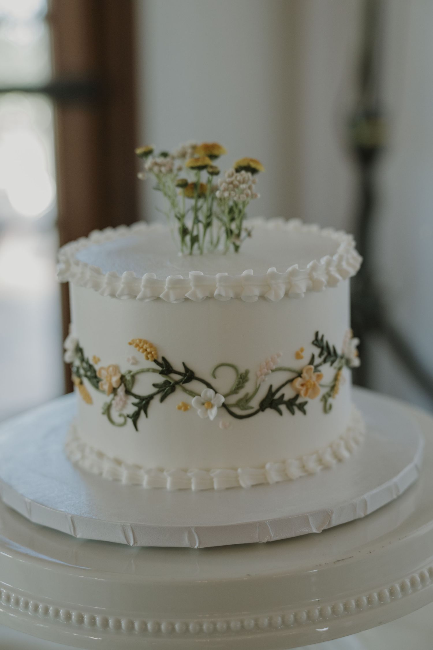 Single-tier white wedding cake with hand-painted wildflower garland, piped pearl border, and fresh daisy topper on white cake stand.