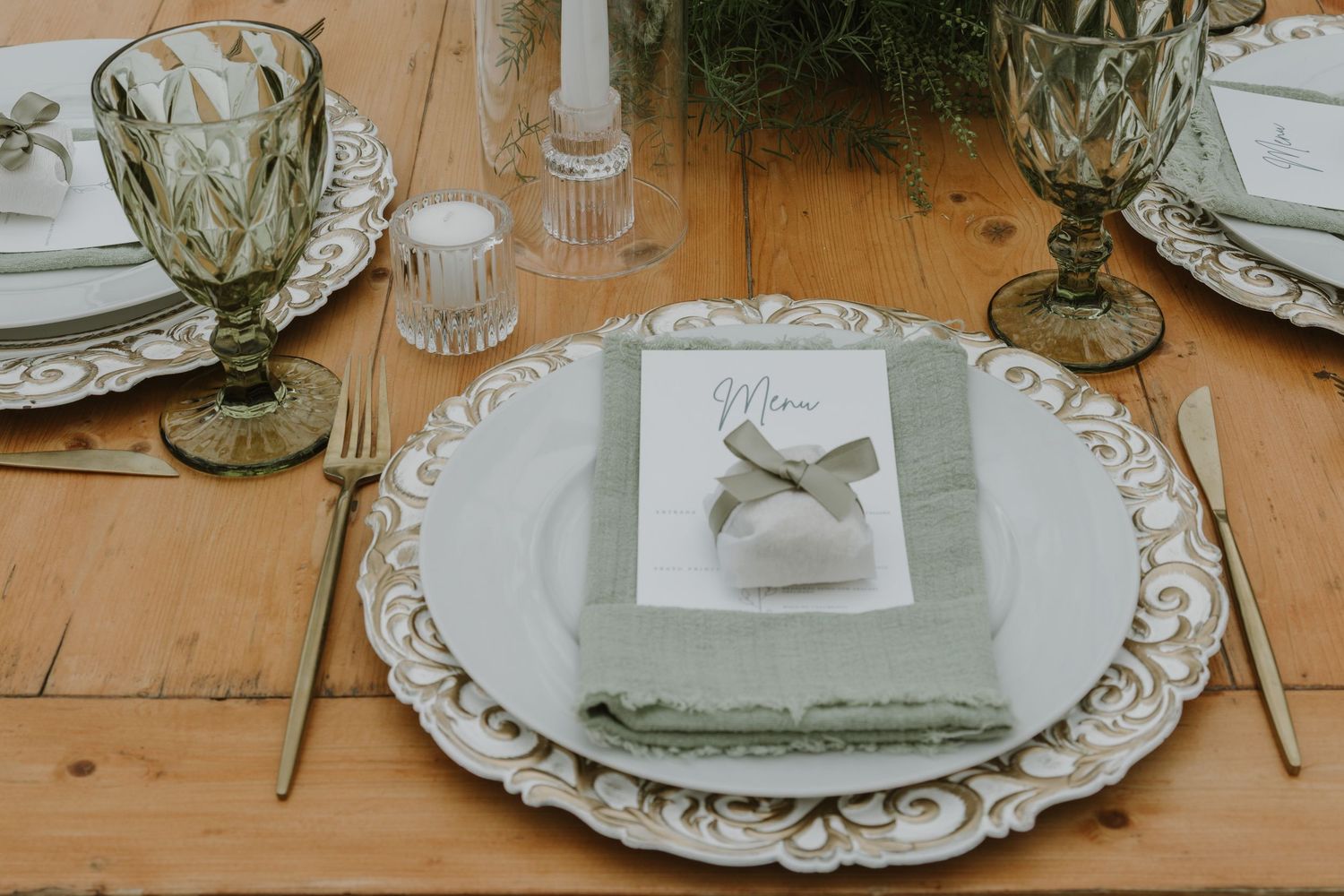 Elegant wedding table setting with sage green napkins, ornate white charger plates, gold cutlery, and green glass goblets.