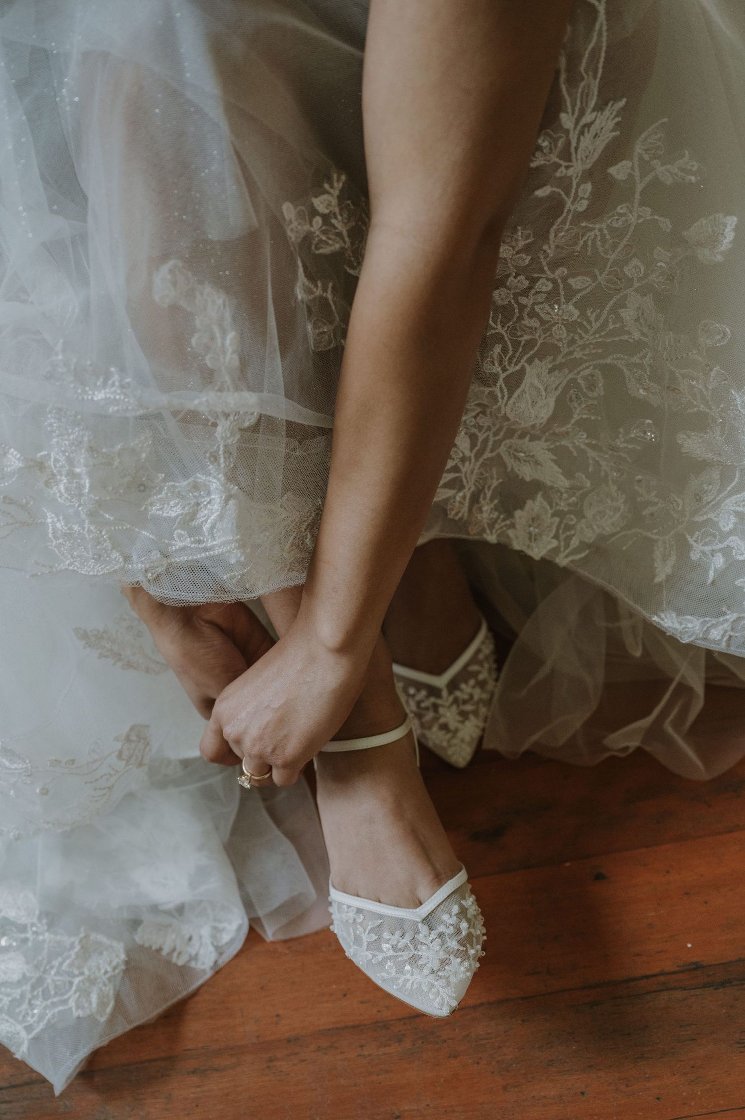 Bride fastening straps of white lace bridal heels while wearing her floral lace wedding gown.