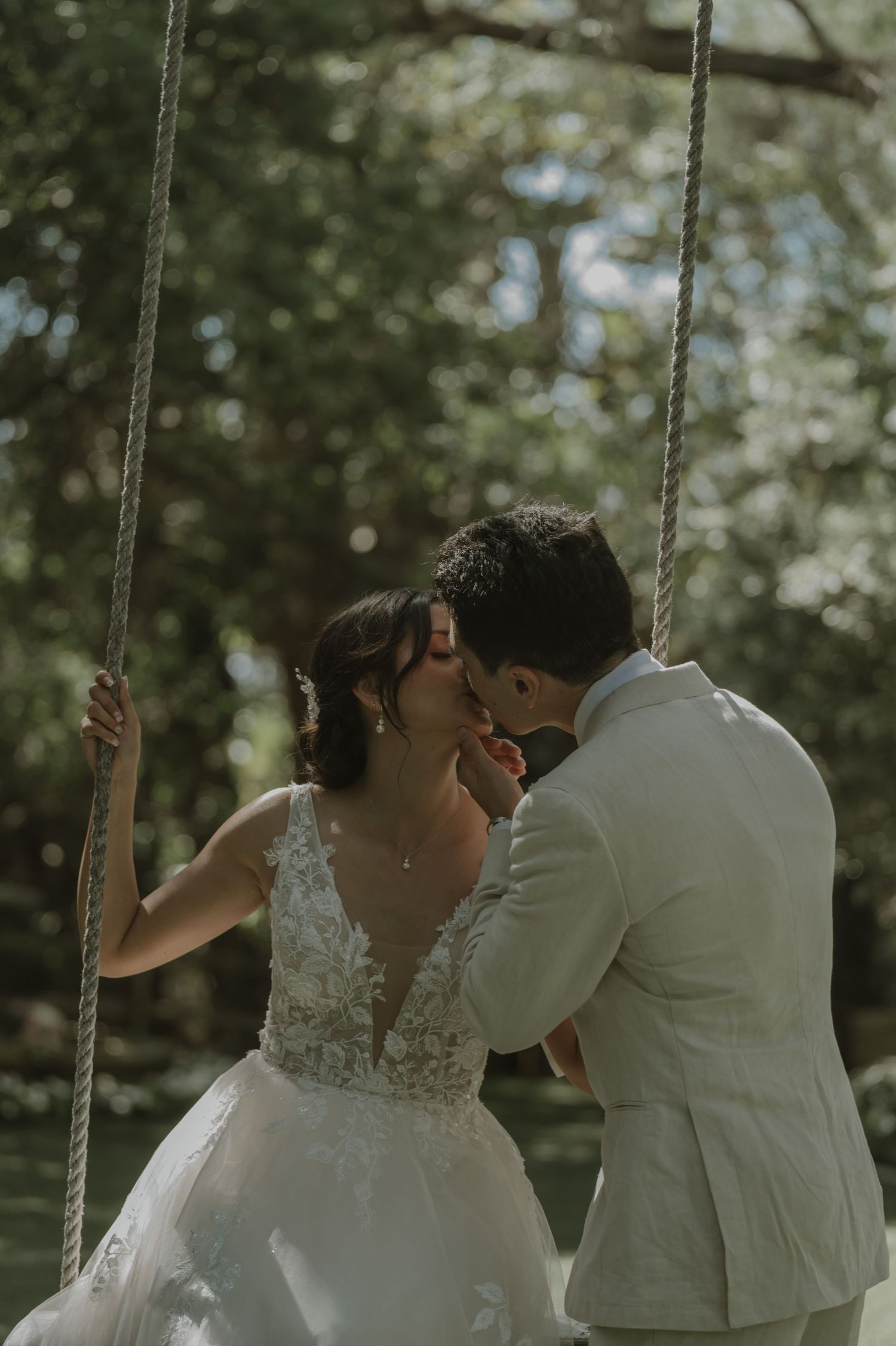 Bride in lace gown and groom in beige suit share a romantic kiss on a rope swing in a sunlit garden.