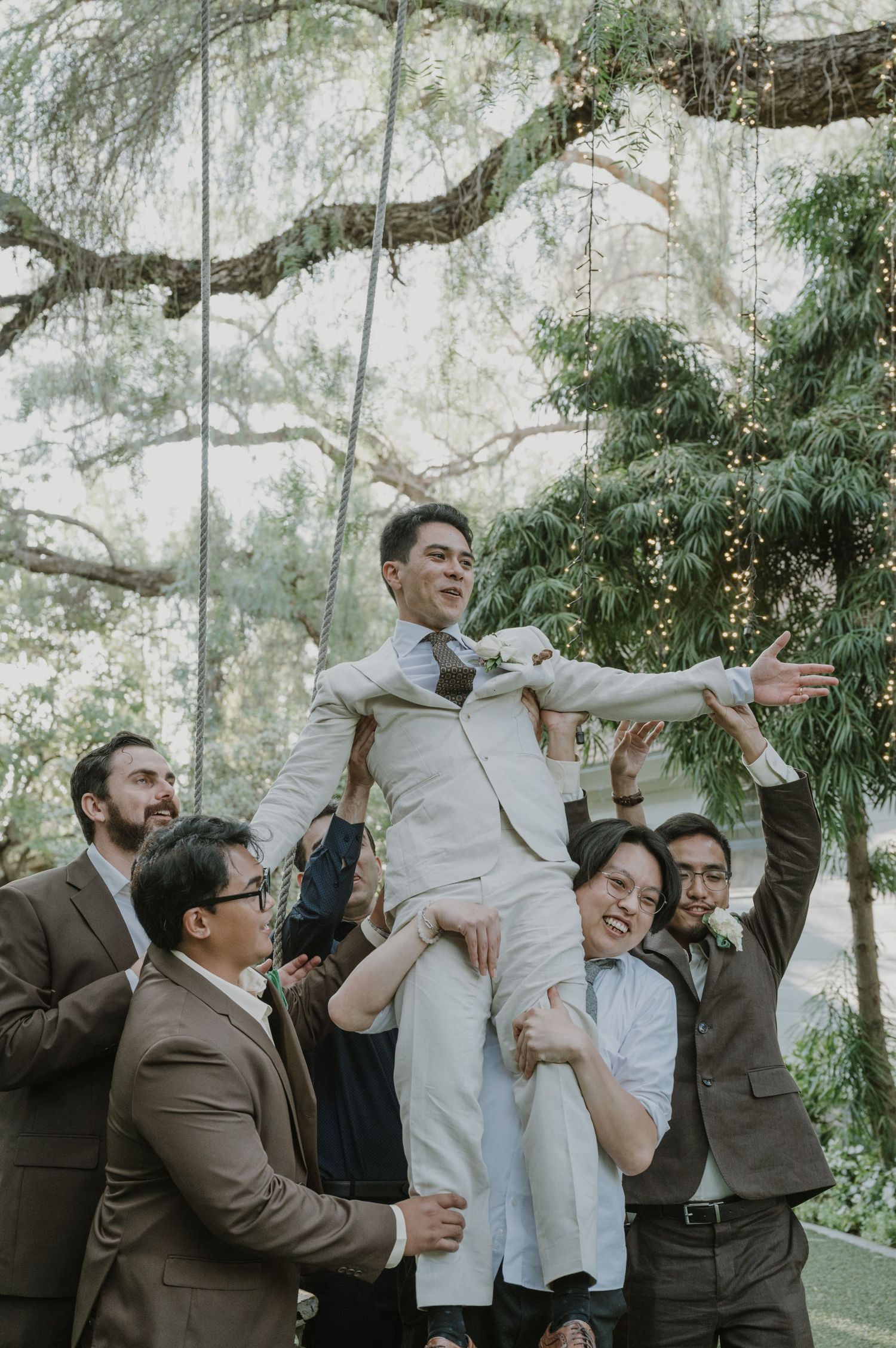 Joyful groom lifted by groomsmen with arms outstretched under fairy-lit trees at wedding.