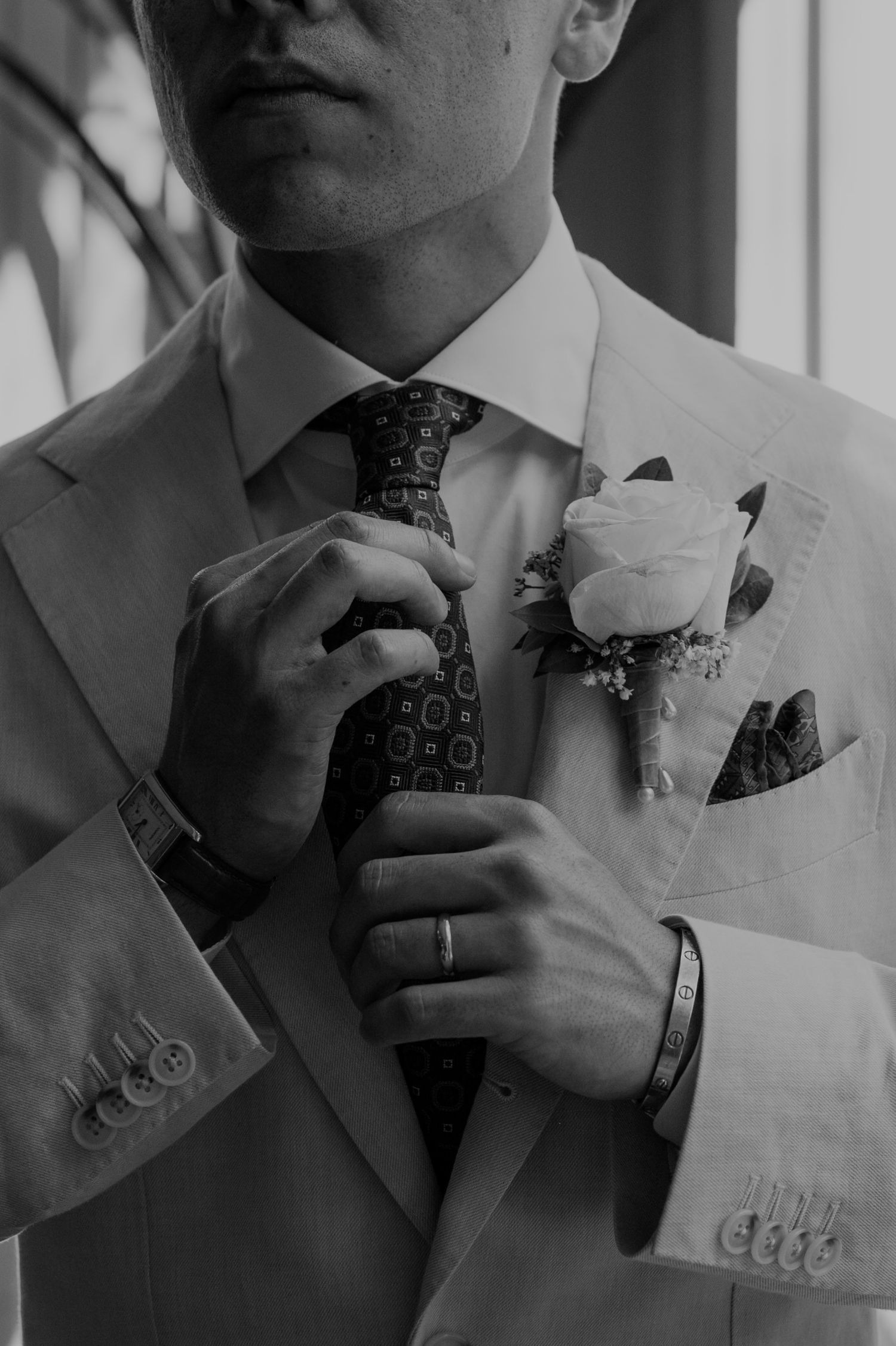 Black and white photo of a groom in a suit adjusting his tie, wearing a boutonniere and ring.