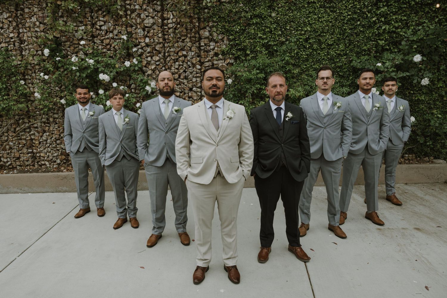 Groom in ivory suit stands before groomsmen in matching gray suits outdoors against a lush green ivy wall.